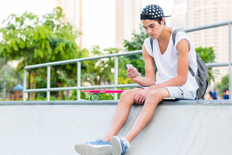 Giovane facendo uso di un cellulare mentre sedendosi allo skatepark immagine stock Giovane facendo uso di un cellulare mentre sedendosi allo skatepark immagine stock