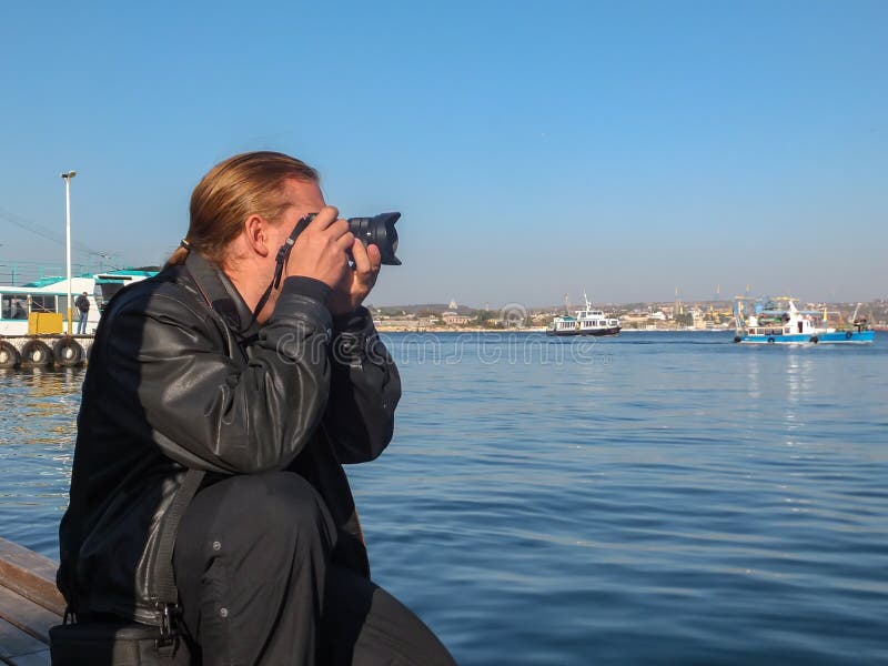 Giovane fotografo bello dell'uomo con capelli lunghi biondi in bomber nero contro la baia blu del mare delle fotografie di Mar Ne fotografia stock Giovane fotografo bello dell'uomo con capelli lunghi biondi in bomber nero contro la baia blu del mare delle fotografie di Mar Ne fotografia stock