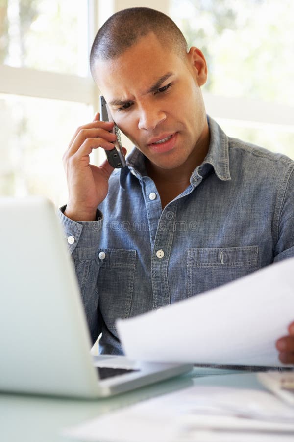 Giovane frustrato sul telefono facendo uso del computer portatile a casa immagini stock Giovane frustrato sul telefono facendo uso del computer portatile a casa immagini stock