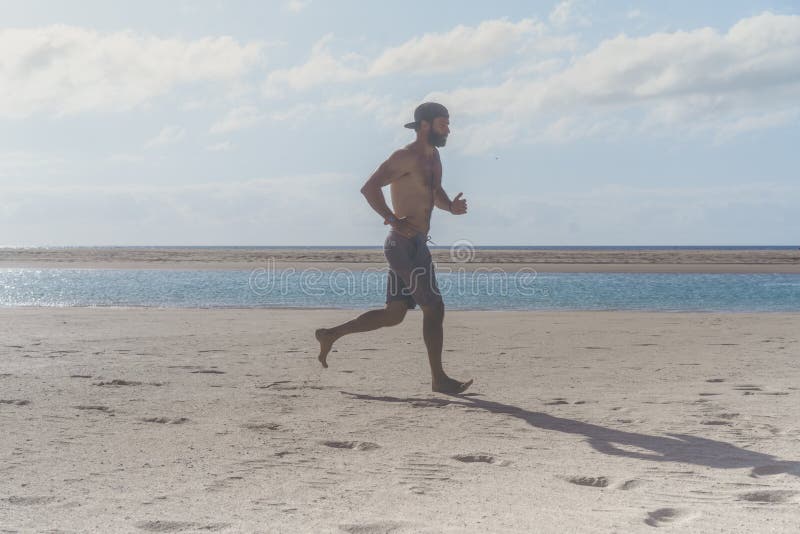 Giovane funzionamento del corridore nella mattina lungo la spiaggia Giovane uomo barbuto che pareggia sulla riva di mare all'alba immagini stock libere da diritti Giovane funzionamento del corridore nella mattina lungo la spiaggia Giovane uomo barbuto che pareggia sulla riva di mare all'alba immagini stock libere da diritti