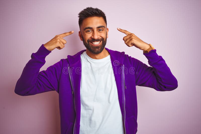 Young indian man wearing purple sweatshirt standing over isolated pink background smiling pointing to head with both hands finger, great idea or thought, good memory. Young indian man wearing purple sweatshirt standing over isolated pink background smiling pointing to head with both hands finger, great idea or thought, good memory