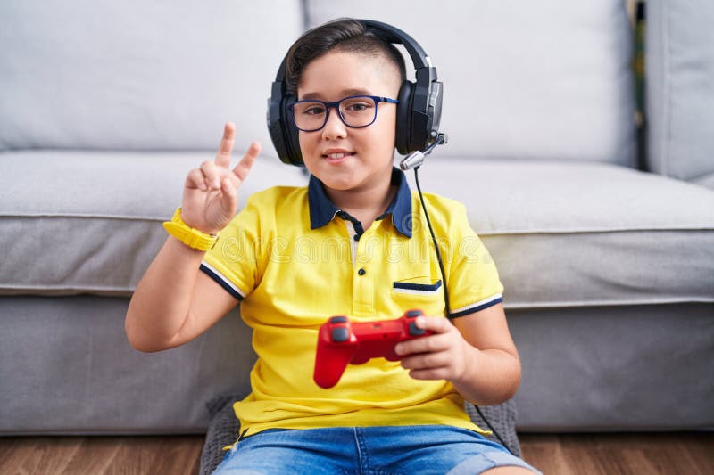 Giovane ispanico che gioca con un videogioco con un controller con cuffie che sorridono con un viso felice mentre fa l'occhiolino immagine stock