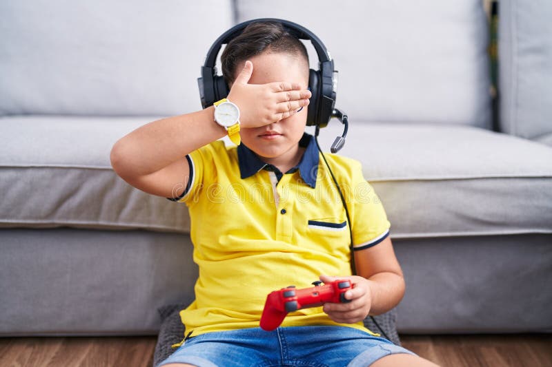 Giovane ispanico che gioca a videogioco con controller che indossa cuffie che coprono gli occhi con la mano che sembrano seri e tr fotografia stock libera da diritti