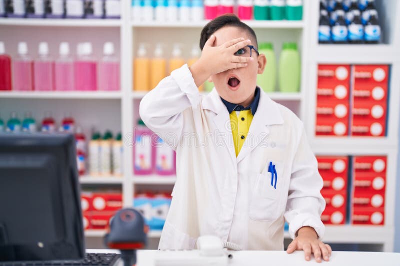 Giovane ispanico che lavora in farmacia drugstore sbirciando sotto shock viso e occhi con la mano che guarda con paura le dita immagine stock libera da diritti