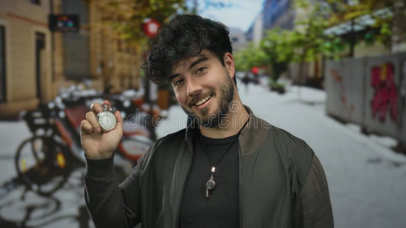 Giovane ispanico con la barba che tiene premuto il cronometro e punta il dito in una strada soleggiata con biciclette e edifici sf fotografie stock libere da diritti