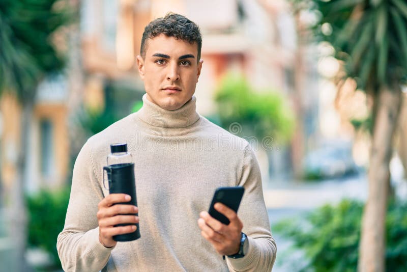 Giovane ispanico con una espressione seria usando smartphone e tenendo in mano una bottiglia d'acqua in città immagini stock libere da diritti