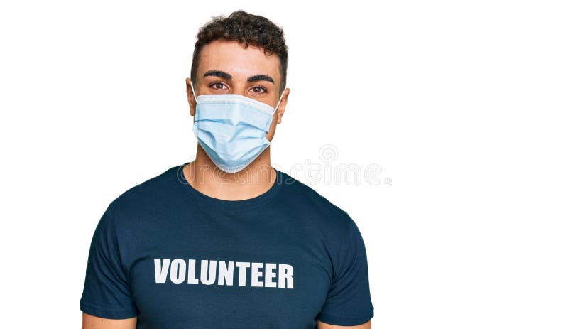 Giovane ispanico con una maschera medica e una maglietta volontaria sembrano positivi e felici e sorridenti con una certa sicurezz fotografie stock