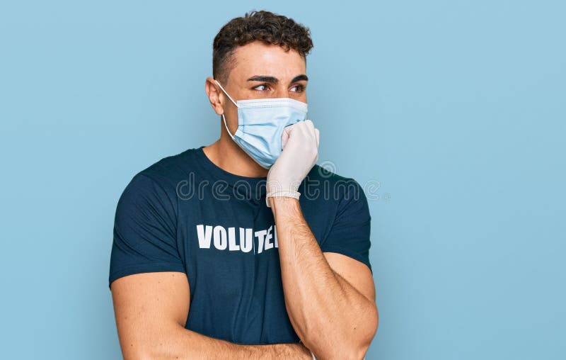 Giovane ispanico indossa una maschera medica e una maglietta volontaria con aspetto stressato e nervoso con mani sulla bocca che p immagini stock libere da diritti