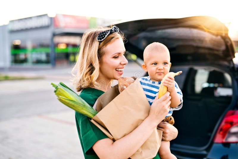 Beautiful young mother with her little baby son in front of a supermarket, holding paper shopping bag. Woman with a boy standing by the car. Beautiful young mother with her little baby son in front of a supermarket, holding paper shopping bag. Woman with a boy standing by the car.