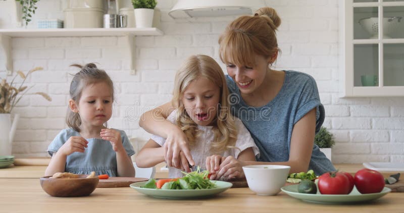 Giovane madre insegna alle figlie a tagliare insalate di verdure in cucina archivi video