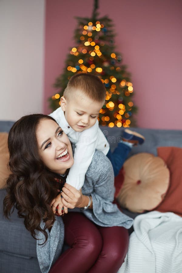 Giovane madre sorridente abbracciando un piccolo figlio felice e posando a casa la vigilia di Natale Ritratto di una giovane madr immagini stock libere da diritti Giovane madre sorridente abbracciando un piccolo figlio felice e posando a casa la vigilia di Natale Ritratto di una giovane madr immagini stock libere da diritti