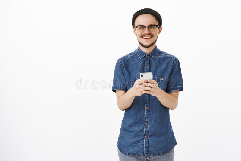 Giovane maschio bello piacevole con la barba e baffi in vetri e smartphone nero della tenuta del beanie che guardano fisso con immagine stock Giovane maschio bello piacevole con la barba e baffi in vetri e smartphone nero della tenuta del beanie che guardano fisso con immagine stock