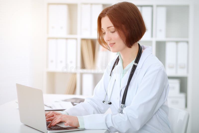 Giovane medico femminile castana che scrive sul comoputer del computer portatile mentre sedendosi alla tavola nell'ufficio dell'o immagini stock libere da diritti Giovane medico femminile castana che scrive sul comoputer del computer portatile mentre sedendosi alla tavola nell'ufficio dell'o immagini stock libere da diritti