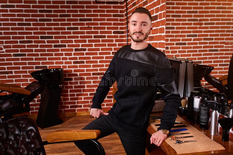 Giovane parrucchiere bello che esamina macchina fotografica mentre stando al parrucchiere ed alle tenute forbici immagine stock libera da diritti Giovane parrucchiere bello che esamina macchina fotografica mentre stando al parrucchiere ed alle tenute forbici immagine stock libera da diritti