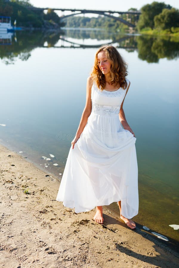 Giovane passeggiata allegra della sposa della ragazza a piedi nudi la costa soleggiata del fiume immagini stock libere da diritti Giovane passeggiata allegra della sposa della ragazza a piedi nudi la costa soleggiata del fiume immagini stock libere da diritti