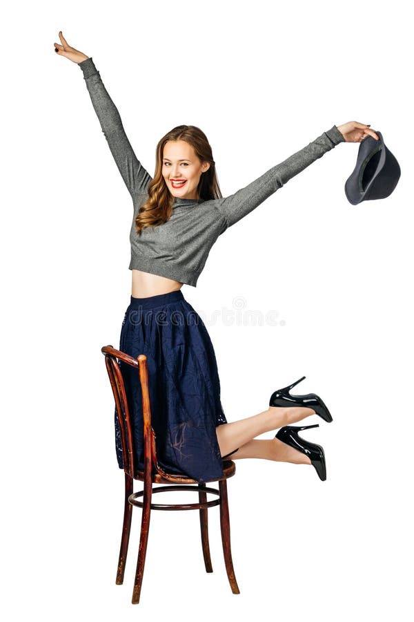 Giovane ragazza allegra con la gonna blu e le ginocchia di maglione grigio sulla sedia e tiene in mano il cappello grigio e ride immagine stock libera da diritti Giovane ragazza allegra con la gonna blu e le ginocchia di maglione grigio sulla sedia e tiene in mano il cappello grigio e ride immagine stock libera da diritti