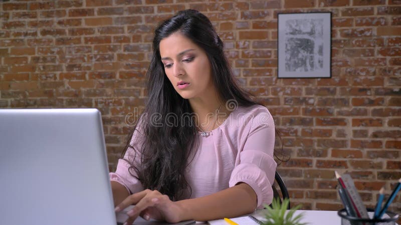 Giovane ragazza araba che lavora nell'ufficio del sottotetto sul computer portatile che scrive, muro di mattoni nel fondo