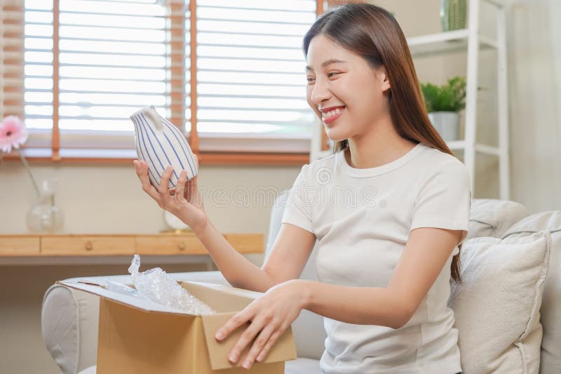 Giovane ragazza asiatica felice, eccitata, seduta sul divano all'apertura di casa e disimballaggio del cartone di cartone, pacchi