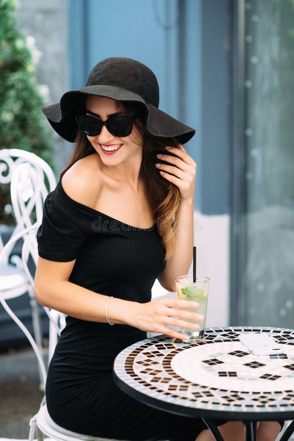 Giovane ragazza graziosa in una seduta black hat in un caffè sulla via e sulla bevanda raffreddata bevente immagini stock Giovane ragazza graziosa in una seduta black hat in un caffè sulla via e sulla bevanda raffreddata bevente immagini stock
