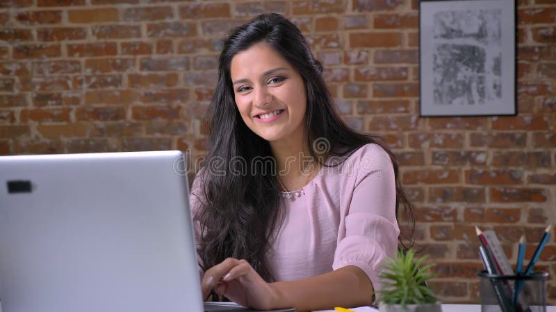 Giovane ragazza seria che lavora nell'ufficio del sottotetto sul computer portatile, scrivente, esaminante macchina fotografica e