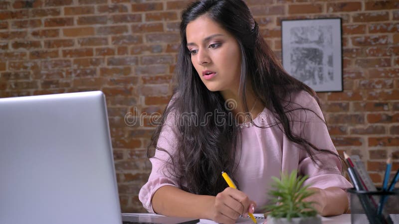 Giovane ragazza seria che lavora in un ufficio del sottotetto su un computer portatile, scrivendo, scrivendo una nota in un taccu