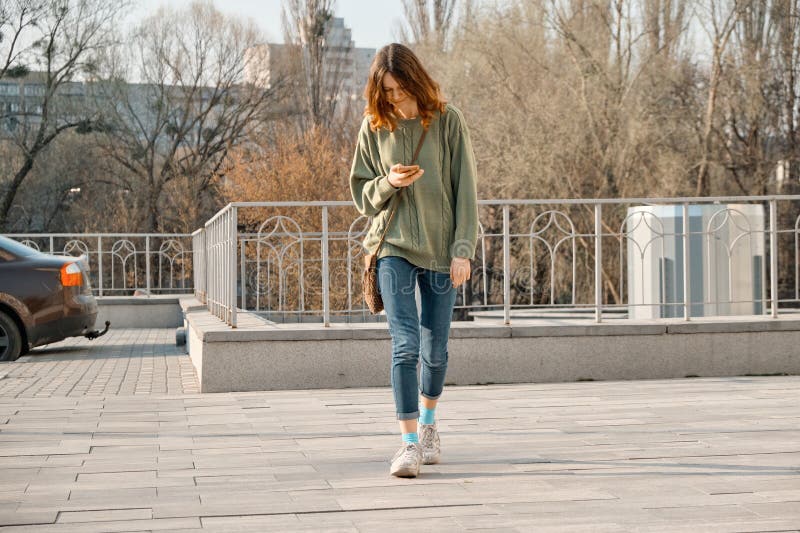 Giovane ragazza teenager che cammina con il telefono, messaggio di testo della lettura sullo smartphone, fondo di giorno soleggia fotografia stock Giovane ragazza teenager che cammina con il telefono, messaggio di testo della lettura sullo smartphone, fondo di giorno soleggia fotografia stock