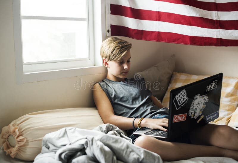 Giovane ragazzo caucasico che si trova facendo uso del computer portatile del computer sul letto fotografie stock libere da diritti Giovane ragazzo caucasico che si trova facendo uso del computer portatile del computer sul letto fotografie stock libere da diritti