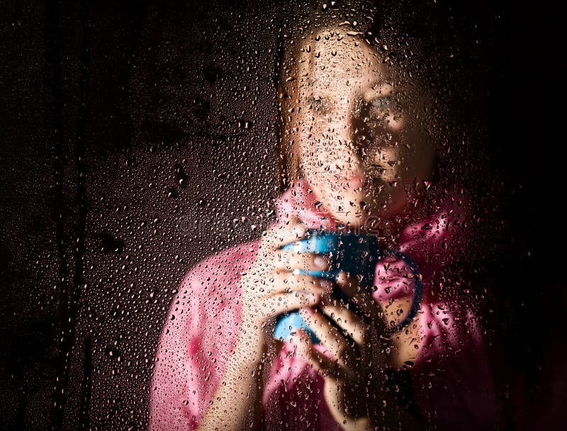 Giovane ritratto triste della donna dietro la finestra nella pioggia con le gocce di pioggia su Ragazza che tiene una tazza dell fotografia stock libera da diritti Giovane ritratto triste della donna dietro la finestra nella pioggia con le gocce di pioggia su Ragazza che tiene una tazza dell fotografia stock libera da diritti
