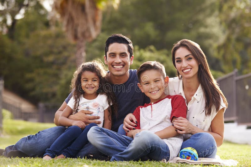 Giovane seduta ispana felice della famiglia sull'erba nel parco che sorride alla macchina fotografica, fine su fotografia stock