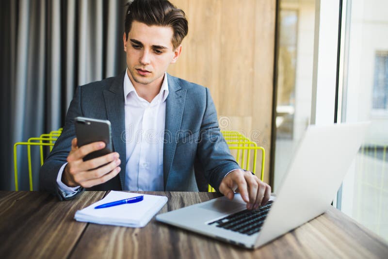 Giovane sicuro in Smart Phone astuto della tenuta di abbigliamento casual ed esaminarlo mentre sedendosi al suo posto di lavoro i immagine stock Giovane sicuro in Smart Phone astuto della tenuta di abbigliamento casual ed esaminarlo mentre sedendosi al suo posto di lavoro i immagine stock
