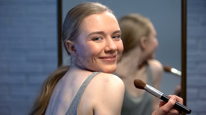 Giovane signora alla moda graziosa che applica la parte anteriore della cipria dello specchio che sorride sulla macchina fotograf fotografie stock Giovane signora alla moda graziosa che applica la parte anteriore della cipria dello specchio che sorride sulla macchina fotograf fotografie stock