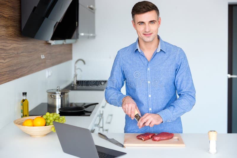 Giovane sorridente attraente che cucina carne sulla cucina immagine stock libera da diritti Giovane sorridente attraente che cucina carne sulla cucina immagine stock libera da diritti