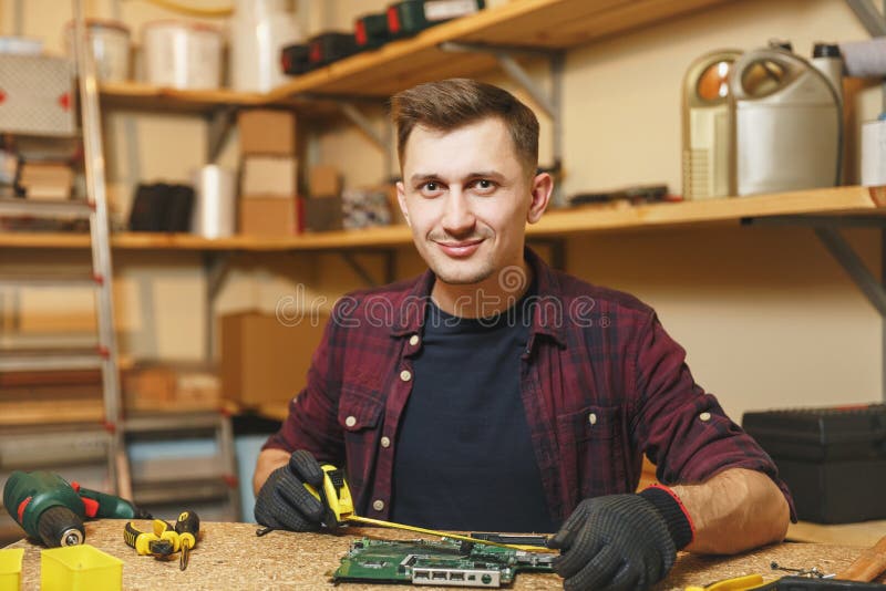 Giovane sorridente bello che lavora nell'officina di carpenteria al posto di legno della tavola con il pezzo di legno immagine stock Giovane sorridente bello che lavora nell'officina di carpenteria al posto di legno della tavola con il pezzo di legno immagine stock