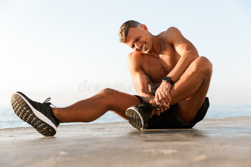 Giovane sportivo senza camicia che soffre da un dolore della caviglia fotografia stock Giovane sportivo senza camicia che soffre da un dolore della caviglia fotografia stock