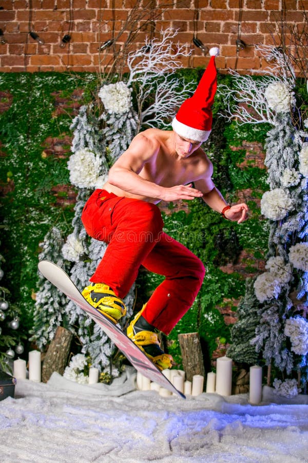 Giovane giovane sportivo sexy in pantaloni rossi e cappello di Santa che saltano con uno snowboard fotografie stock libere da diritti Giovane giovane sportivo sexy in pantaloni rossi e cappello di Santa che saltano con uno snowboard fotografie stock libere da diritti