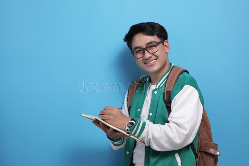 Giovane studente asiatico di baseball con una giacca verde di baseball e scritto su un libro fotografia stock Giovane studente asiatico di baseball con una giacca verde di baseball e scritto su un libro fotografia stock