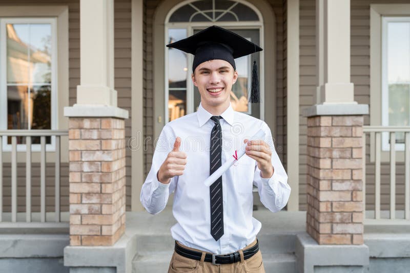 Giovane studente maschio che tiene un diploma mentre indossando un cappuccio di graduazione davanti ad una casa mentre tenendo po fotografia stock libera da diritti Giovane studente maschio che tiene un diploma mentre indossando un cappuccio di graduazione davanti ad una casa mentre tenendo po fotografia stock libera da diritti