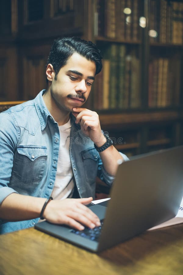Giovane uomo arabo astuto che lavora nella biblioteca con il computer immagine stock Giovane uomo arabo astuto che lavora nella biblioteca con il computer immagine stock