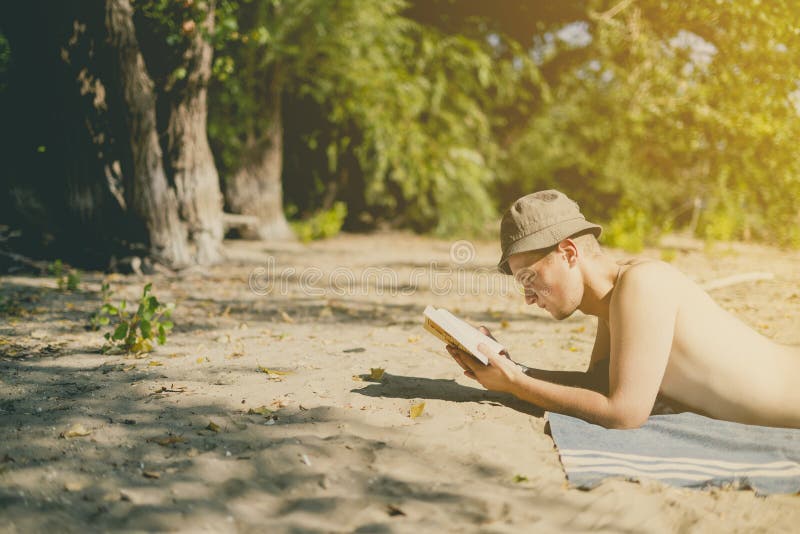 Giovane uomo attraente bello nel Panama che si trova sulla spiaggia sabbiosa di cielo e che legge un libro un giorno di estate immagini stock libere da diritti Giovane uomo attraente bello nel Panama che si trova sulla spiaggia sabbiosa di cielo e che legge un libro un giorno di estate immagini stock libere da diritti
