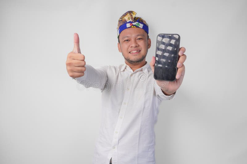 Giovane uomo balinese indossando una camicia bianca e un copricapo tradizionale presentando uno schermo vuoto sul suo telefono men immagini stock