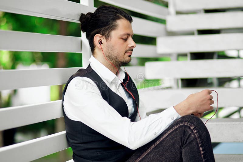 Giovane uomo barbuto bello dei pantaloni a vita bassa che utilizza smartphone e musica d'ascolto con le cuffie nella città fotografia stock libera da diritti Giovane uomo barbuto bello dei pantaloni a vita bassa che utilizza smartphone e musica d'ascolto con le cuffie nella città fotografia stock libera da diritti