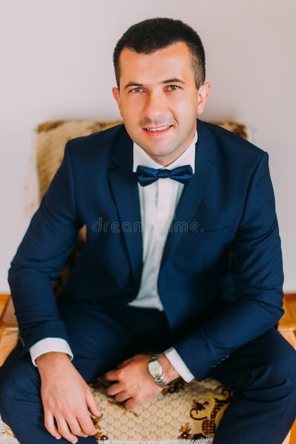 Giovane uomo bello alla moda e sicuro in vestito blu scuro con il farfallino che si siede nella sedia d'annata contro il fondo gr fotografia stock libera da diritti Giovane uomo bello alla moda e sicuro in vestito blu scuro con il farfallino che si siede nella sedia d'annata contro il fondo gr fotografia stock libera da diritti