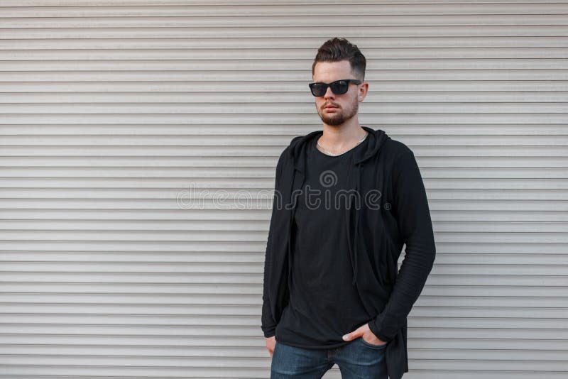 Giovane uomo bello brutale dei pantaloni a vita bassa con una barba in una maglietta alla moda in una maglia con cappuccio nera a fotografia stock Giovane uomo bello brutale dei pantaloni a vita bassa con una barba in una maglietta alla moda in una maglia con cappuccio nera a fotografia stock