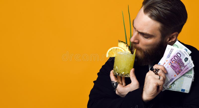 Giovane uomo d'affari felice con vetro del cocktail in vestiti convenzionali che tengono mazzo di banconote dei soldi fotografia stock libera da diritti Giovane uomo d'affari felice con vetro del cocktail in vestiti convenzionali che tengono mazzo di banconote dei soldi fotografia stock libera da diritti