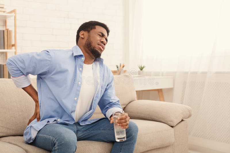 Young african-american man with back pain, pressing on hip with painful expression, sitting on sofa at home with glass of water, copy space. Young african-american man with back pain, pressing on hip with painful expression, sitting on sofa at home with glass of water, copy space