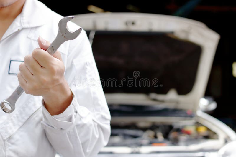 Giovane uomo professionale del meccanico in chiave uniforme della tenuta con il cappuccio aperto al garage di riparazione Concett fotografie stock Giovane uomo professionale del meccanico in chiave uniforme della tenuta con il cappuccio aperto al garage di riparazione Concett fotografie stock