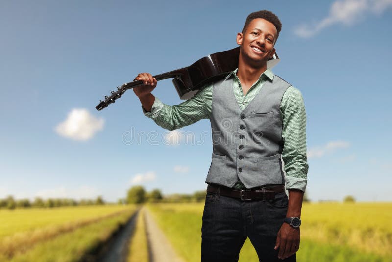 Giovane uomo sorridente dei pantaloni a vita bassa che posa allegro con la chitarra sulla spalla sul fondo vago del paesaggio fotografie stock libere da diritti Giovane uomo sorridente dei pantaloni a vita bassa che posa allegro con la chitarra sulla spalla sul fondo vago del paesaggio fotografie stock libere da diritti