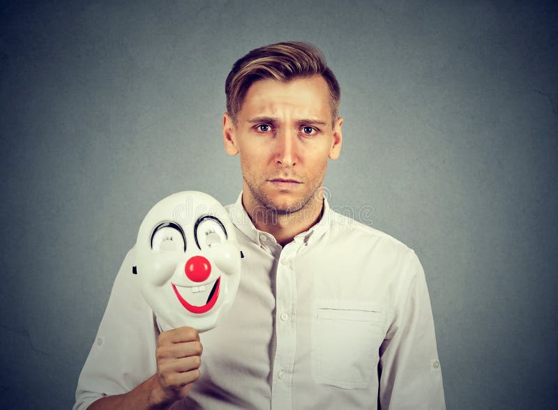 Giovane uomo triste con la maschera felice del pagliaccio fotografia stock libera da diritti Giovane uomo triste con la maschera felice del pagliaccio fotografia stock libera da diritti