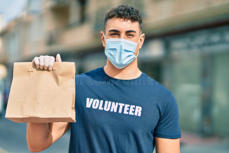 Giovane volontario ispanico indossa una maschera medica che porta il sacchetto di consegna in città immagini stock