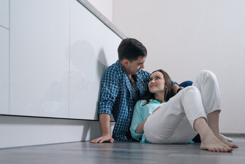 Giovani coppie amorose sul pavimento di parquet nella seduta interna della cucina bianca e sorridere ad a vicenda immagine stock libera da diritti Giovani coppie amorose sul pavimento di parquet nella seduta interna della cucina bianca e sorridere ad a vicenda immagine stock libera da diritti